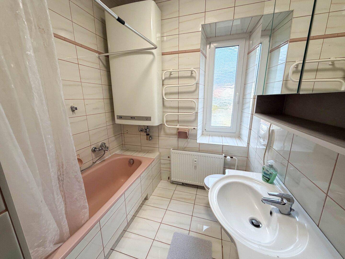 Badezimmer mit Badewanne, Waschbecken und Fenster, ausgestattet mit hellen Fliesen und Heizkörper.