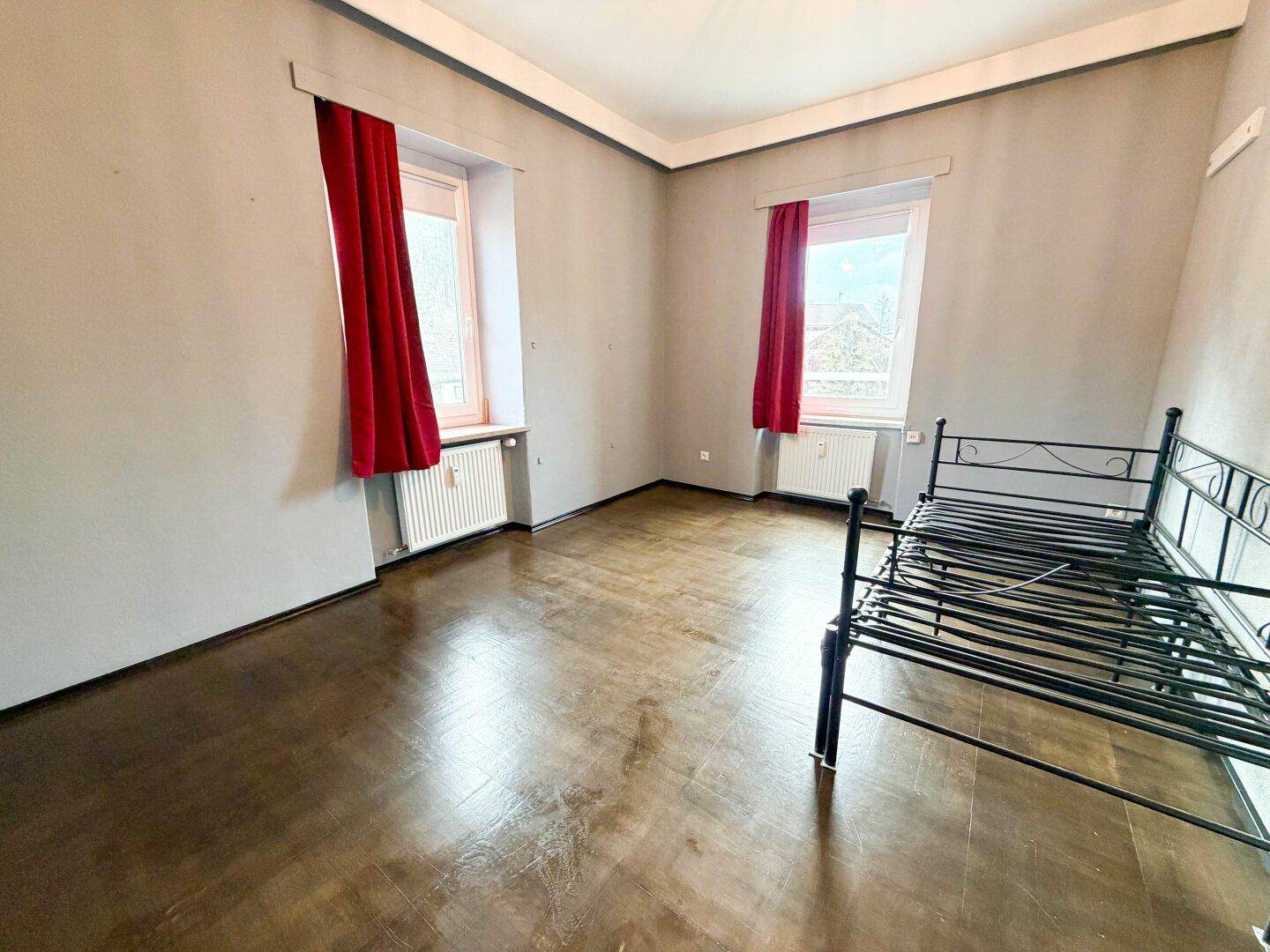 Schlafzimmer mit Parkettboden, roten Vorhängen und zwei Fenstern, bietet viel Tageslicht.
