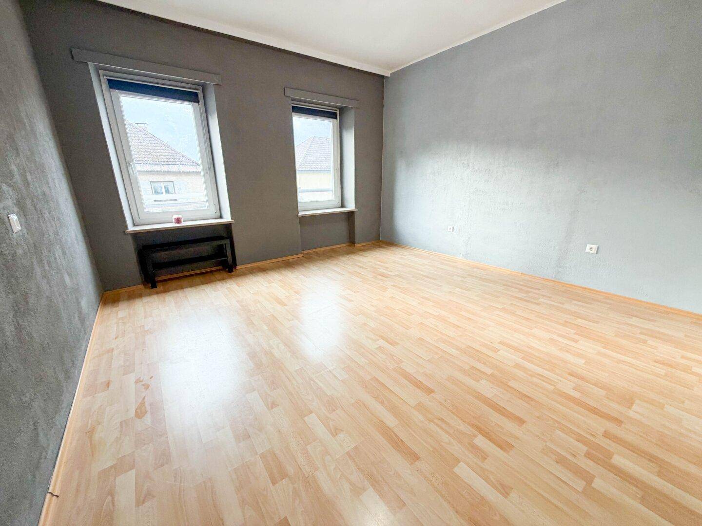 Helles Zimmer mit Laminatboden und grauen Wänden, ausgestattet mit zwei Fenstern.
