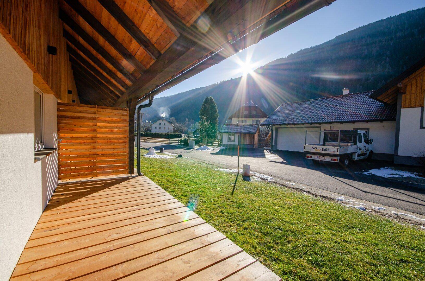 Überdachte Holzterrasse mit Blick auf die Straße, weitere Häuser und die umliegende Berglandschaft.