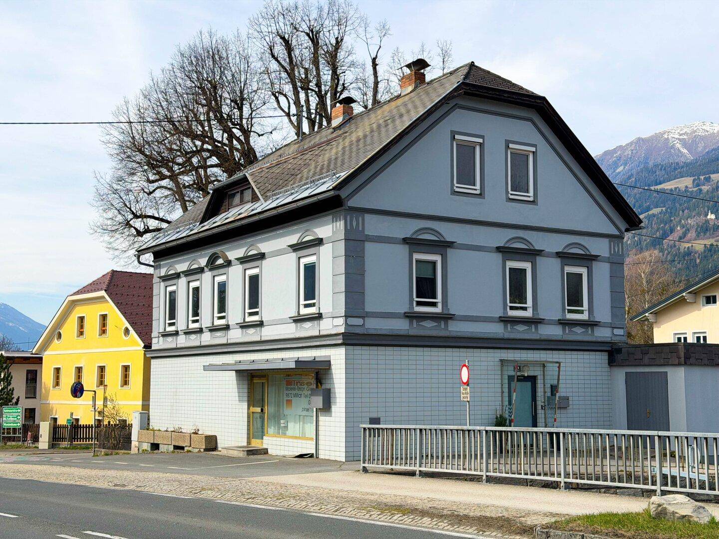 Ansicht des historischen Gebäudes mit grauer Fassade, Mansarddach und Fenstern, umgeben von Natur.