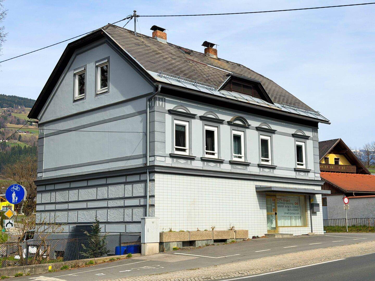 Seitenansicht des Gebäudes mit grauer Fassade, Mansarddach und Blick auf die umliegende Landschaft.