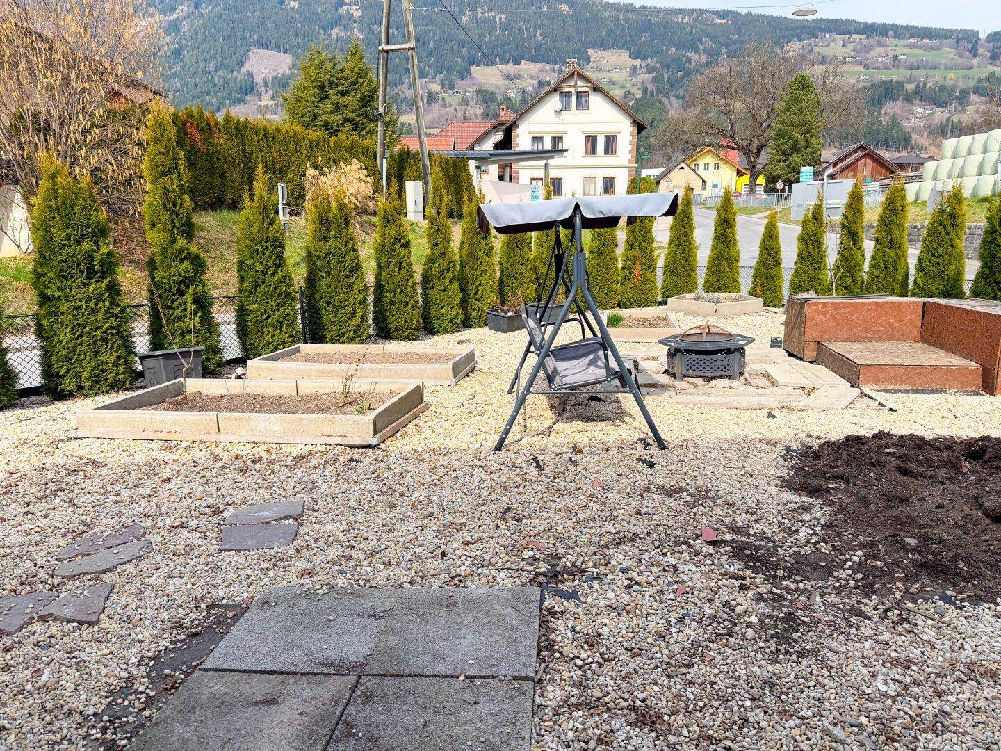 Gartenbereich mit Schaukel, Feuerstelle und erhöhten Beeten, umgeben von Hecken und Bäumen.