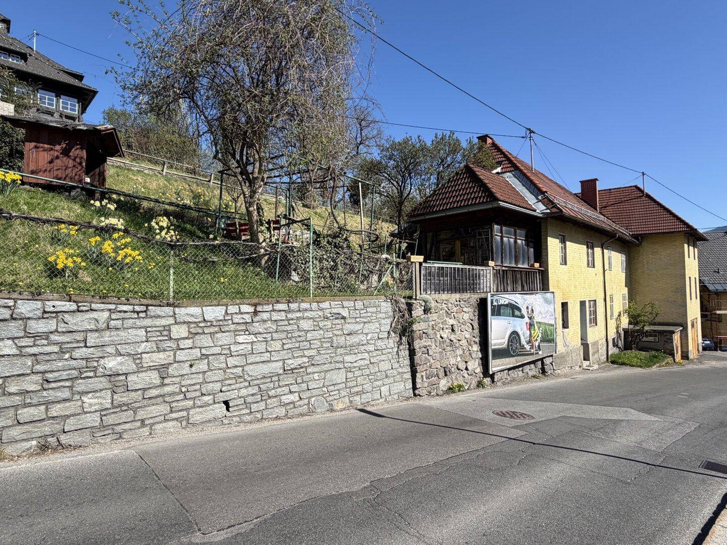 Ansicht des Gebäudes von der Straße, mit einer Steinmauer und blühenden Narzissen im Vorgarten.