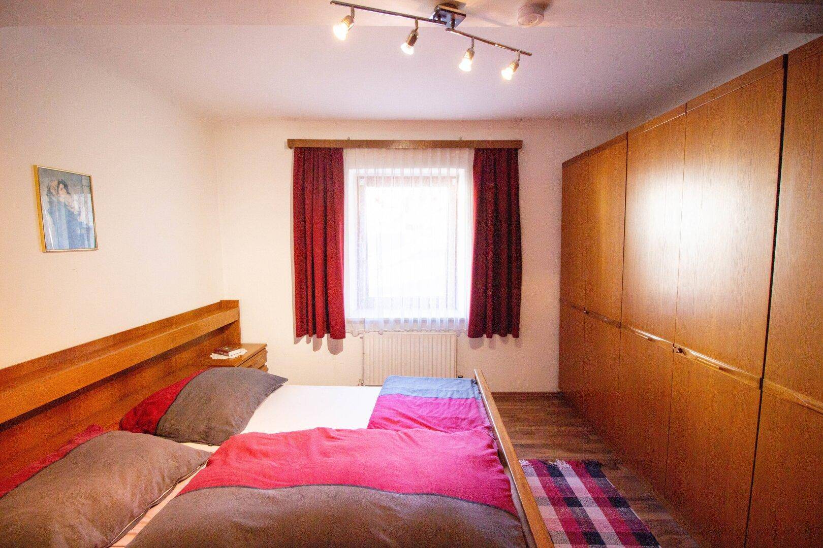 Gemütliches Schlafzimmer mit Holzbett, Einbauschränken und Fenster mit roten Vorhängen.
