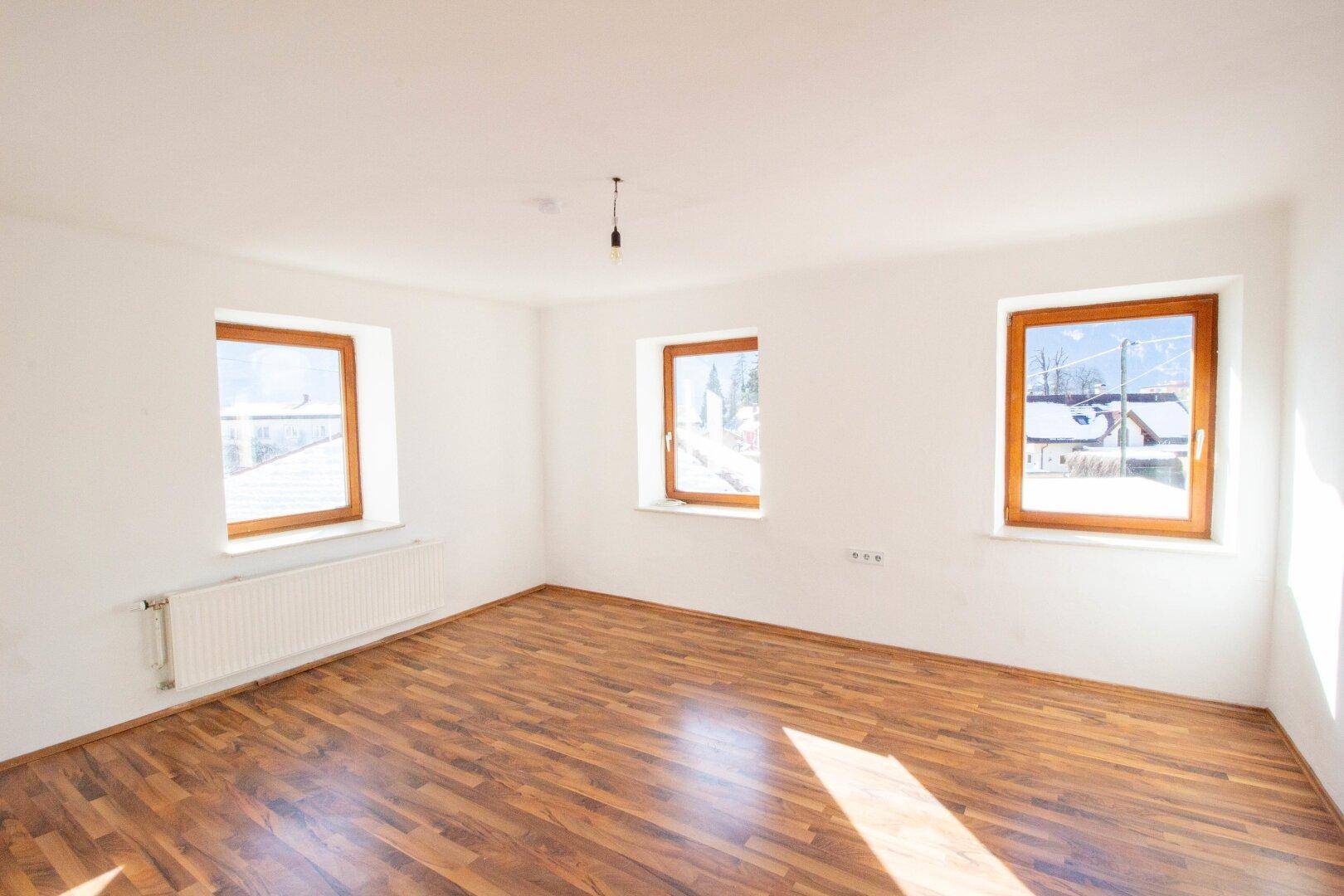 Helles, leeres Zimmer mit Holzboden und drei Fenstern, die einen Blick auf die verschneite Landschaft bieten.