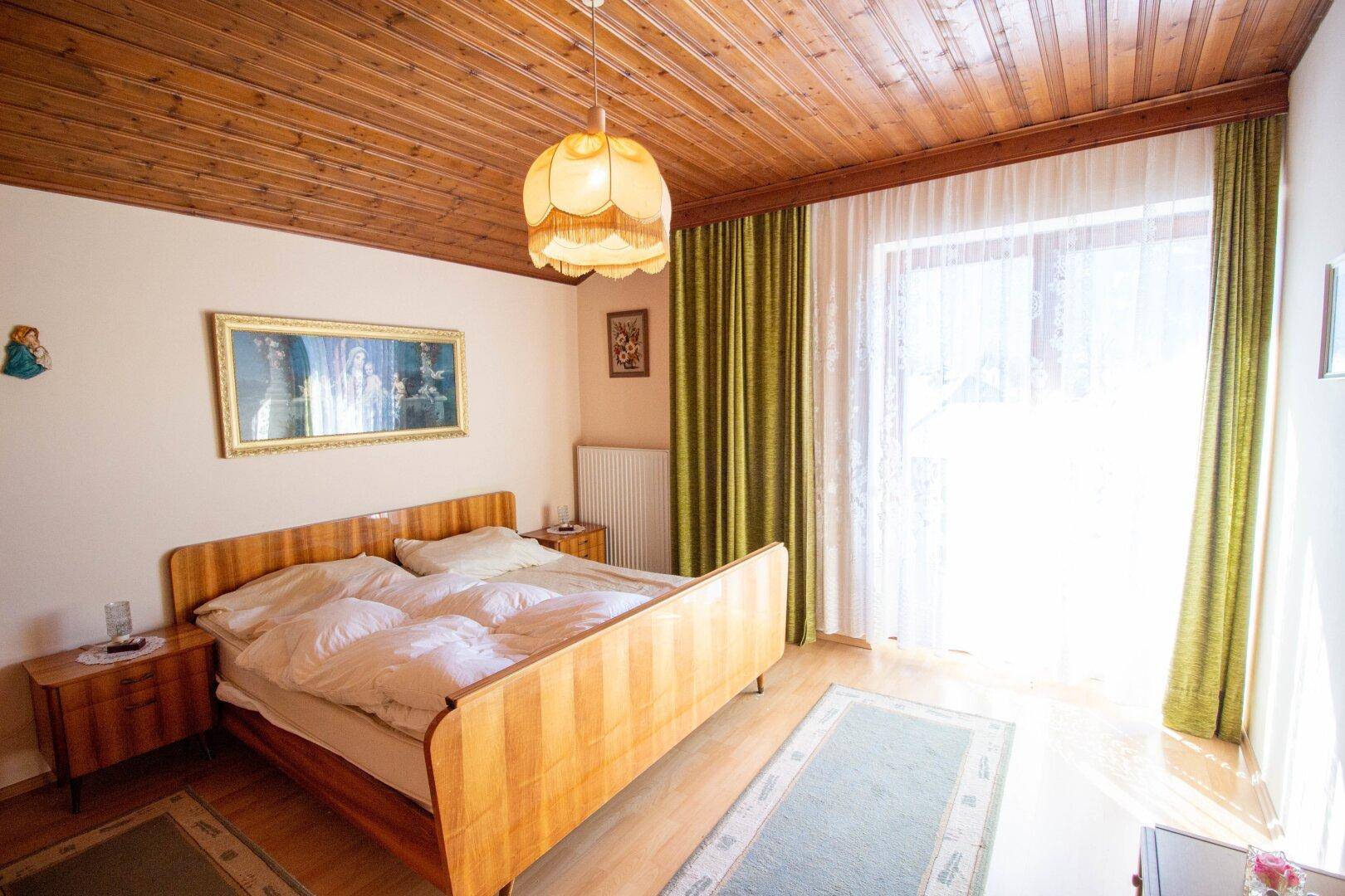 Helles Schlafzimmer mit Holzbett, Holzdecke und großem Fenster mit Zugang nach draußen.