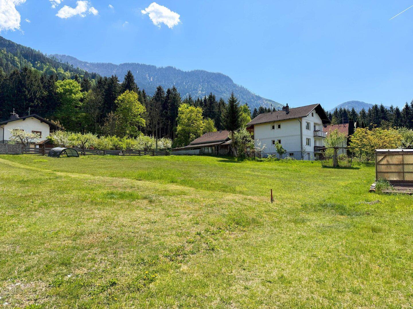 Sonnige Wiese mit Blick auf bewaldete Berge und einige Häuser in ländlicher Umgebung.