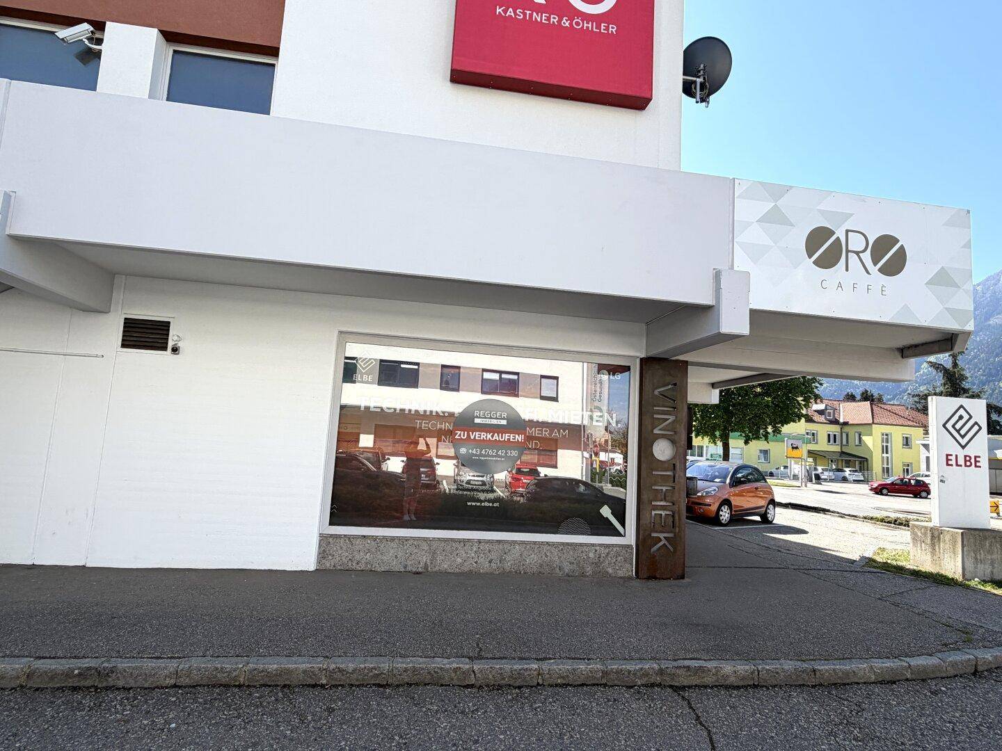 Fassadenansicht eines Geschäfts mit Schaufensterwerbung und einem "ORO CAFFE" Schild, im Hintergrund sind Berge zu sehen.