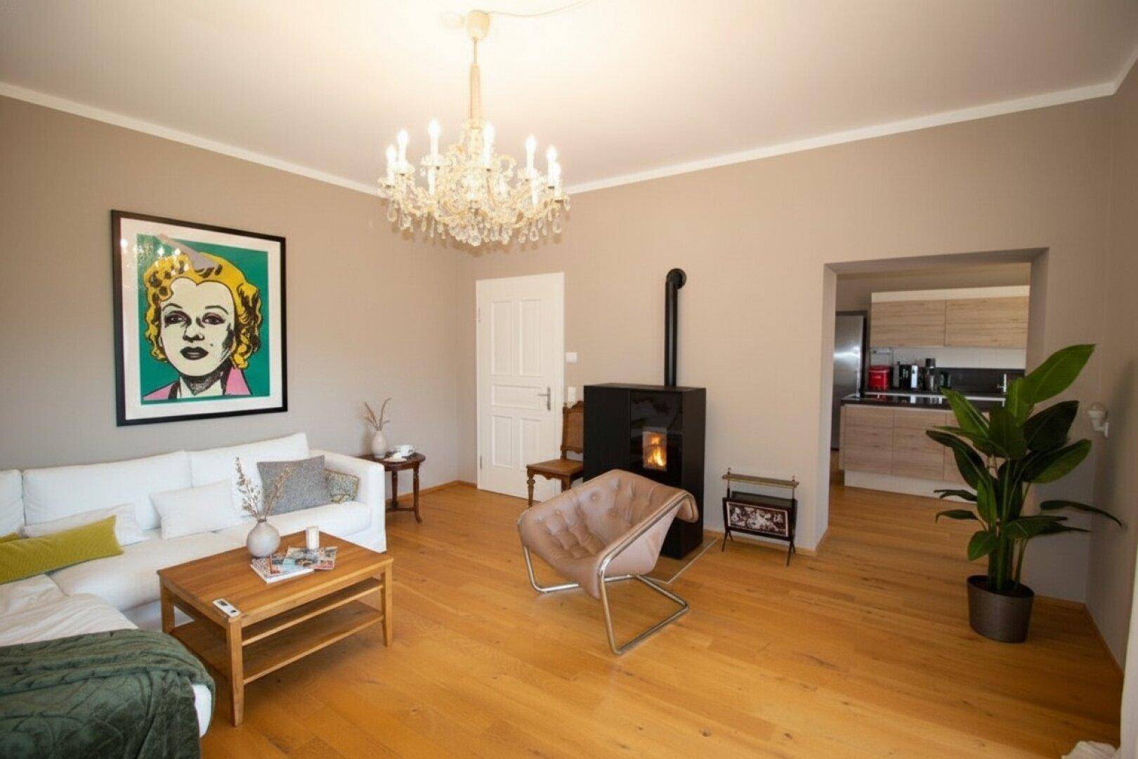 Helles Wohnzimmer mit Holzboden, Kaminofen und Zugang zu einer modernen Küche.