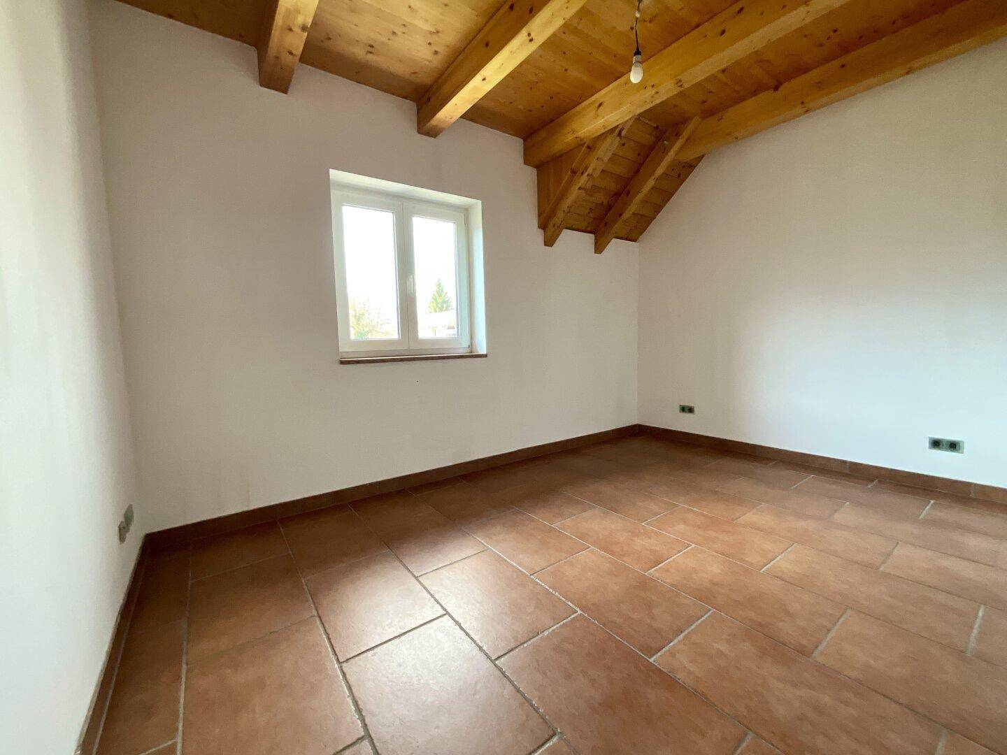 Unmöbliertes Schlafzimmer mit Terrakottafliesen, schräger Holzbalkendecke und einem hellen Fenster.