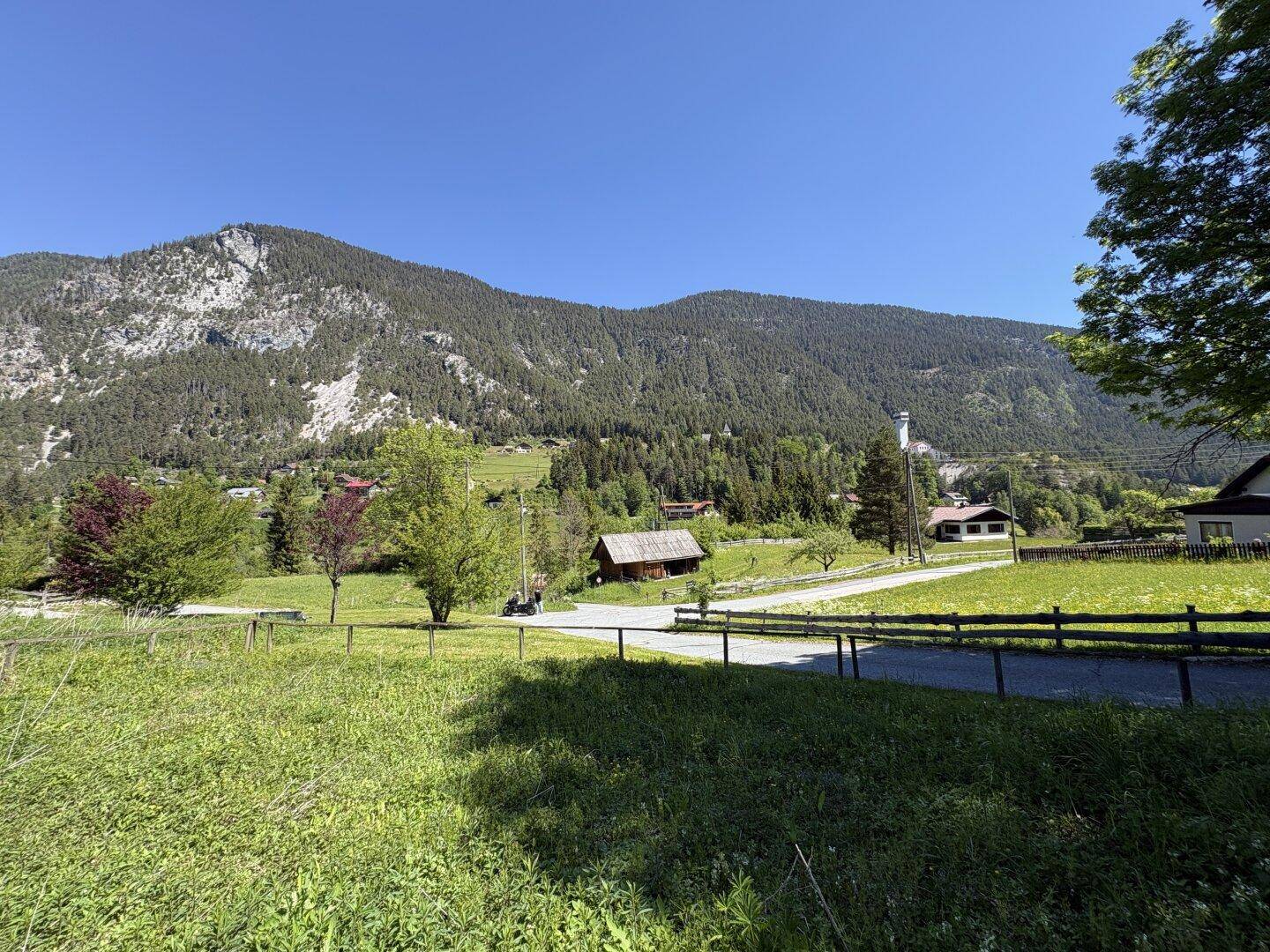 Weitläufige Landschaft mit mehreren Häusern, umgeben von grünen Wiesen und majestätischen Bergen unter klarem Himmel.