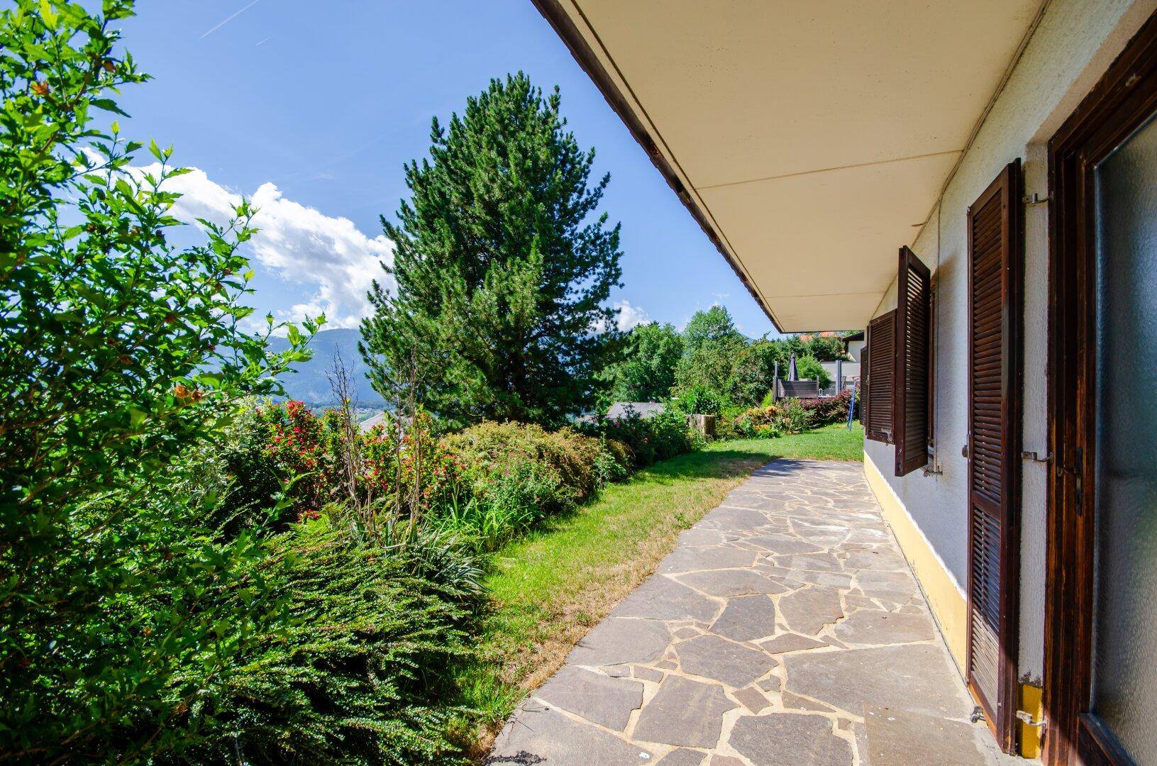 Seitlicher Gartenbereich mit Steinplattenweg, üppiger Bepflanzung und Blick auf die Berge.