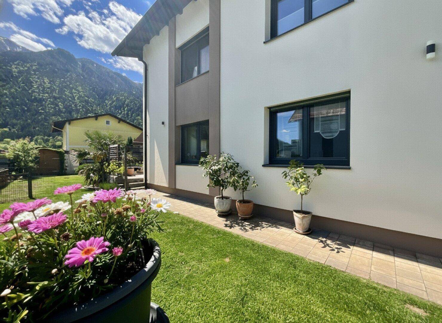 Seitenansicht des Hauses mit gepflegtem Rasen, Blumen und Blick auf die Bergkulisse.