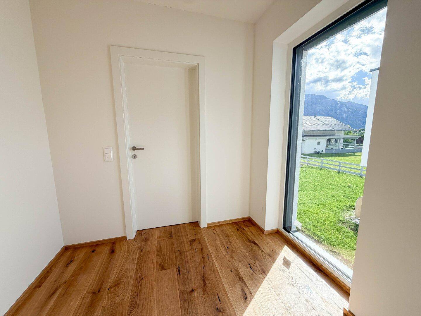 Helles Zimmer mit Holzboden und einem großen Fenster, das einen Blick ins Grüne ermöglicht.
