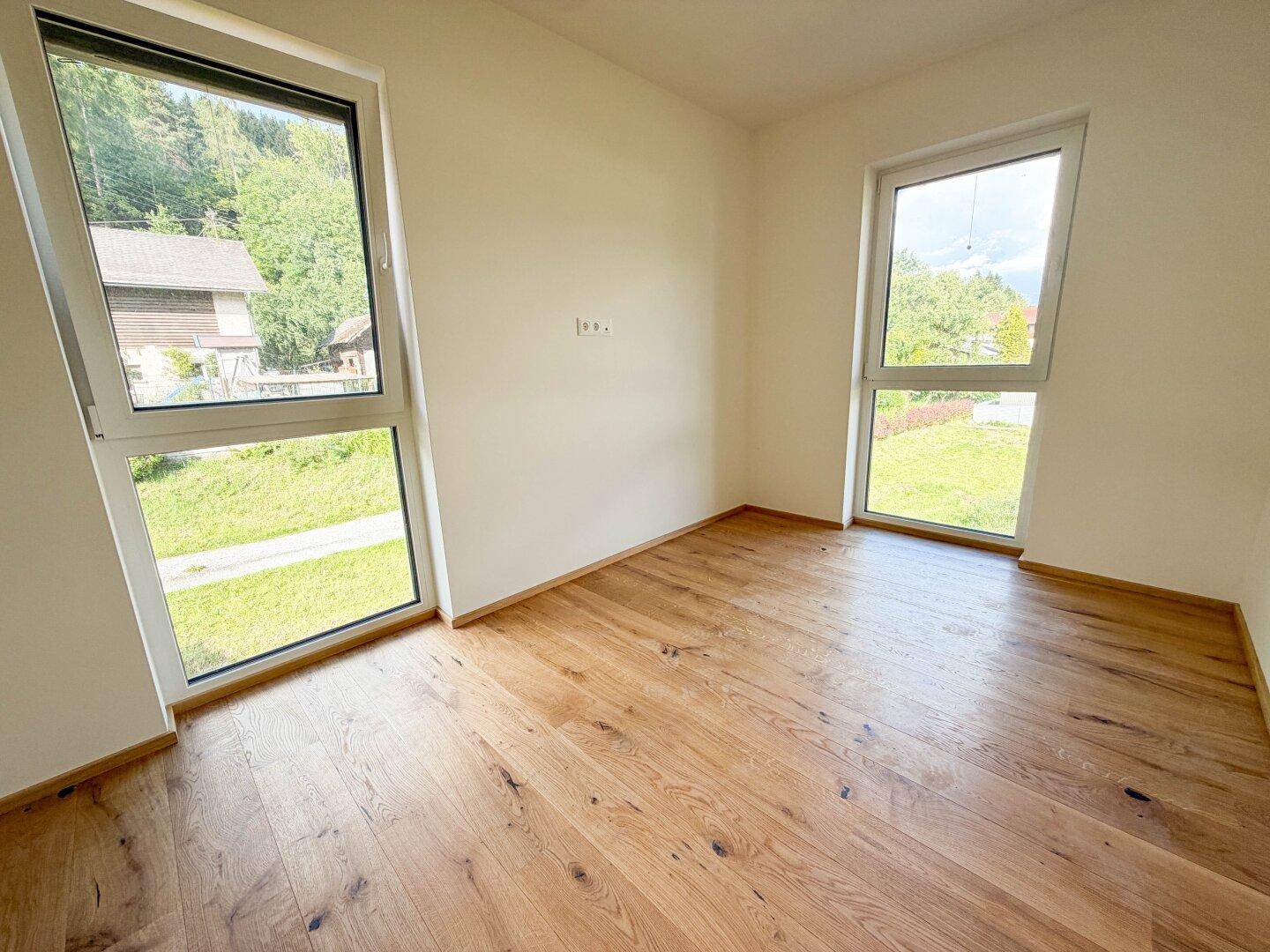 Helles Zimmer mit Holzboden und zwei großen Fenstern, die viel Tageslicht hereinlassen.