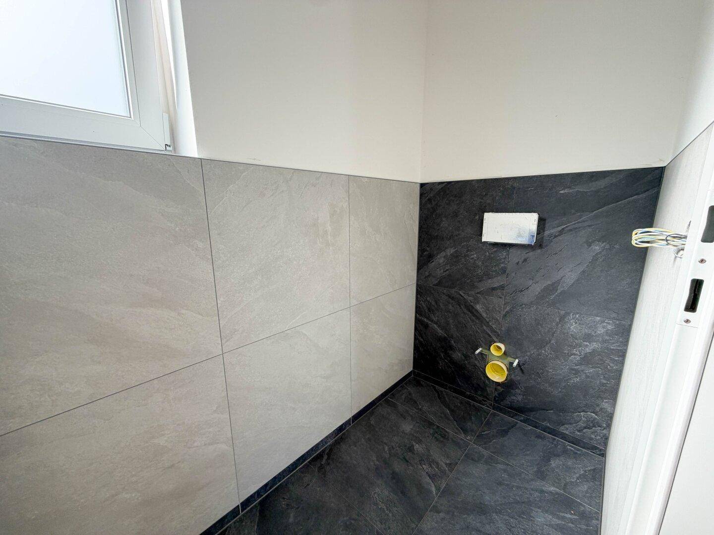 Kleines Gäste-WC mit modernen grauen und schwarzen Fliesen, bereit für die Installation der Sanitäranlagen.