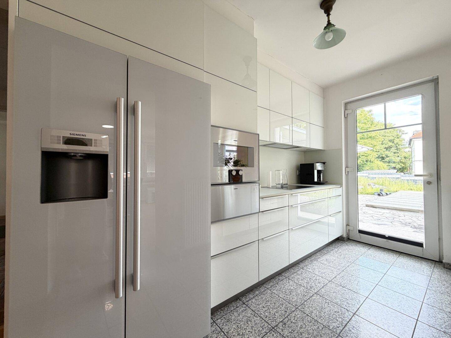 Moderne Küche mit weißen Hochglanzfronten, großem Kühlschrank und Zugang zur Terrasse.