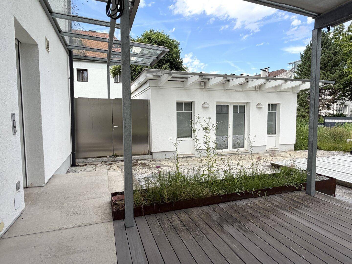 Moderne Holzterrasse mit Glasüberdachung und Blick auf ein weißes Nebengebäude.