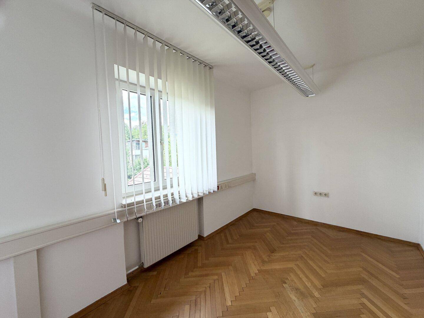 Heller Raum mit klassischem Fischgrätparkett und einem Fenster mit vertikalen Jalousien, das viel Licht spendet.