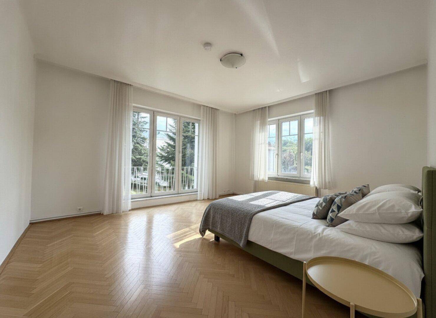 Helles Schlafzimmer mit Fischgrätparkett, großzügigen Fenstern und Blick ins Grüne.