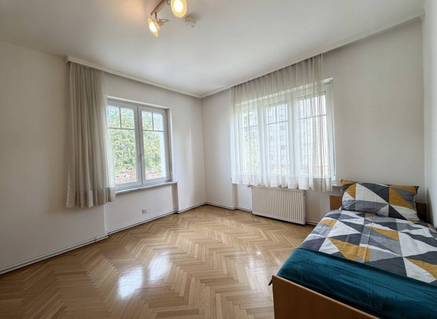 Gemütliches Schlafzimmer mit klassischem Fischgrätparkett und viel Tageslicht durch große Fenster.