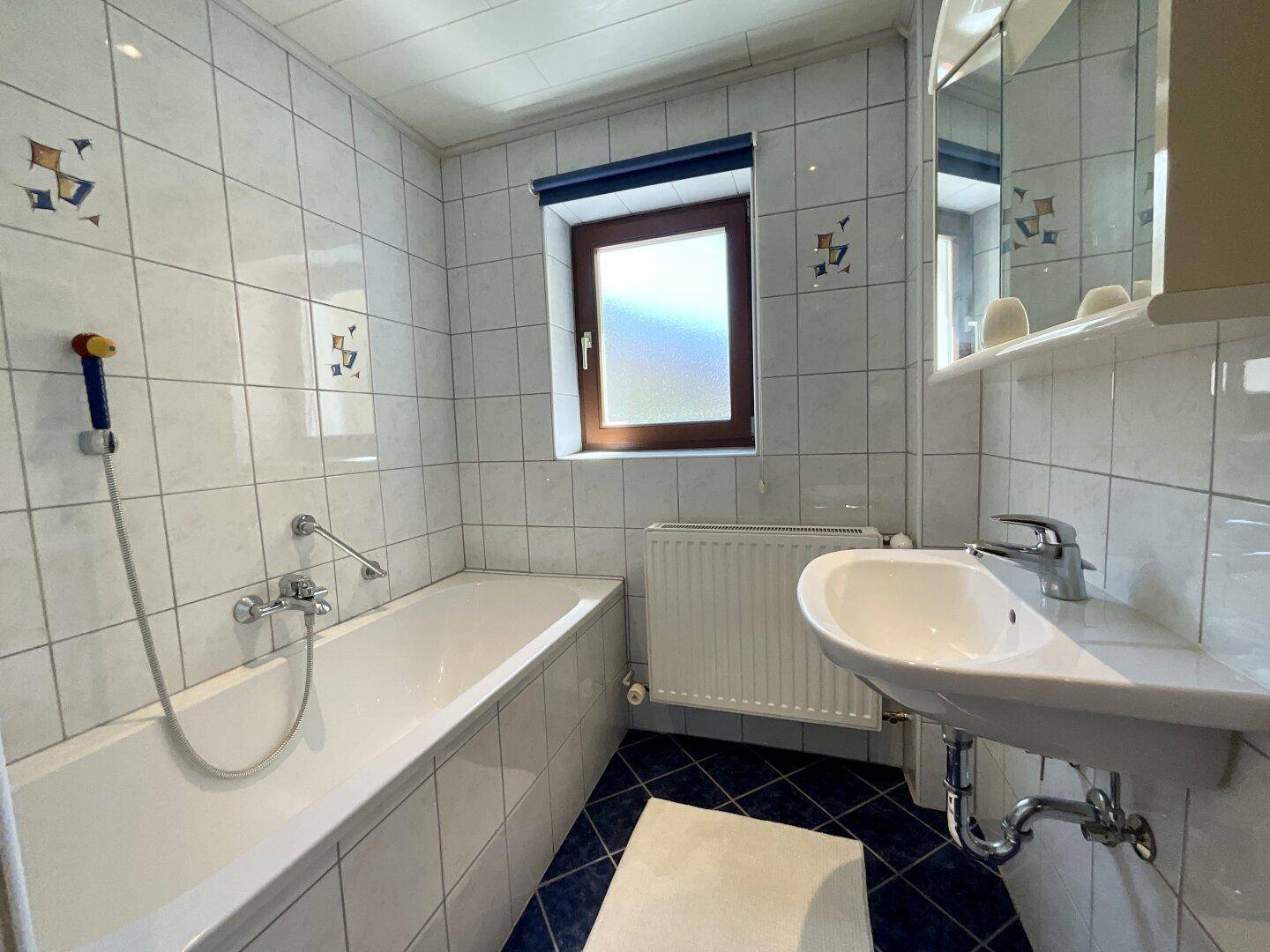 Badezimmer mit Badewanne, Waschbecken und Fenster, ausgestattet mit weißen Fliesen und blauen Akzenten.