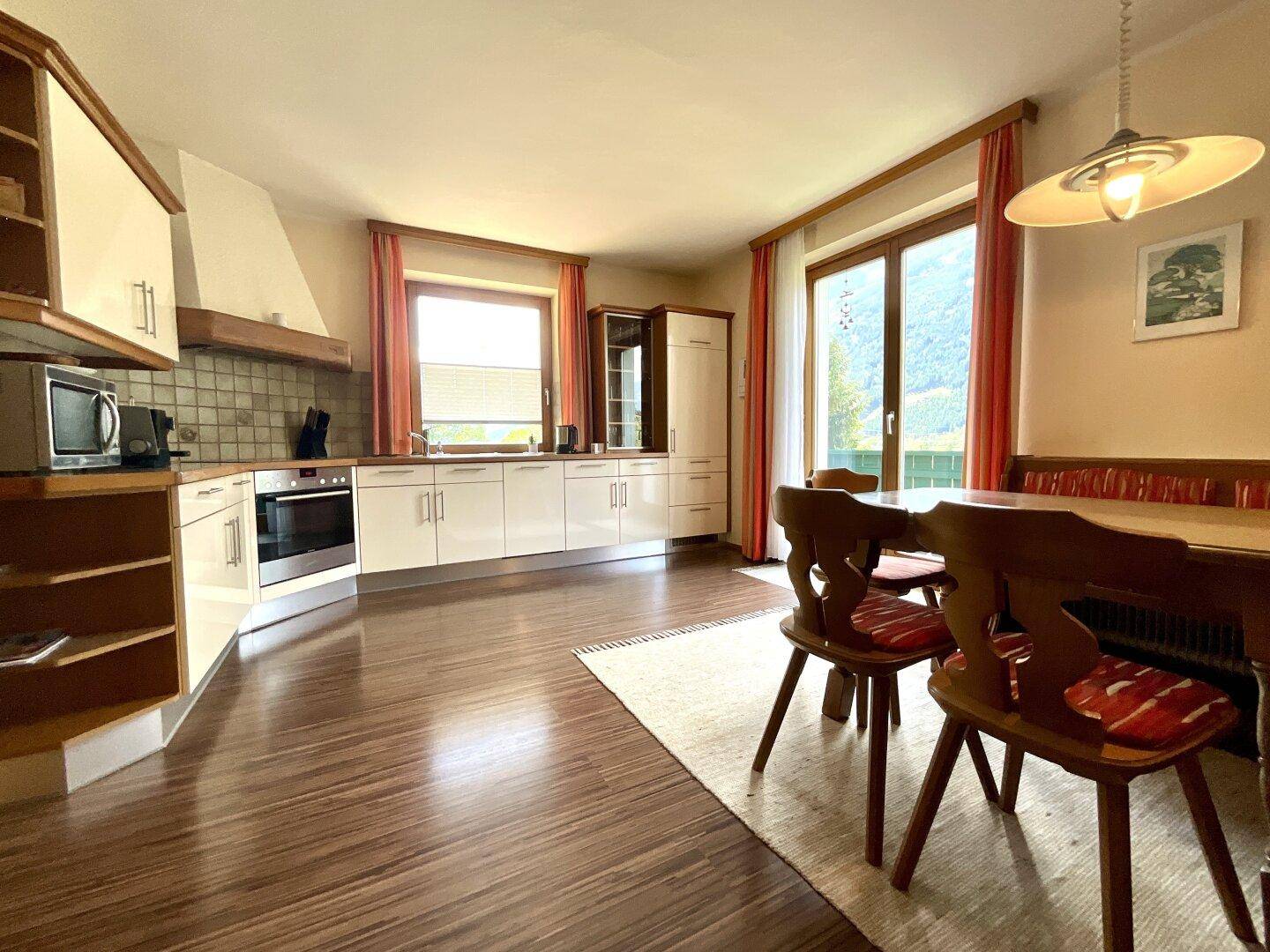 Geräumige Wohnküche mit Holzelementen, Esstisch und Zugang zu einem Balkon mit Bergblick.