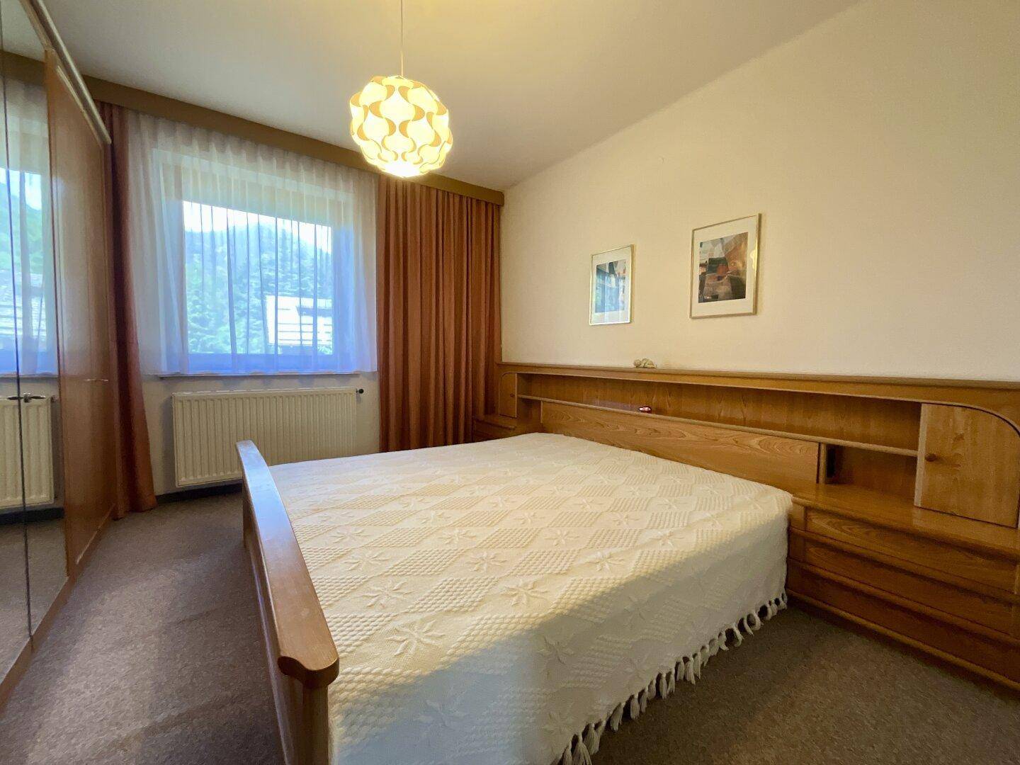 Schlafzimmer mit Doppelbett, Holzmöbeln und großem Fenster, das viel Tageslicht hereinlässt.