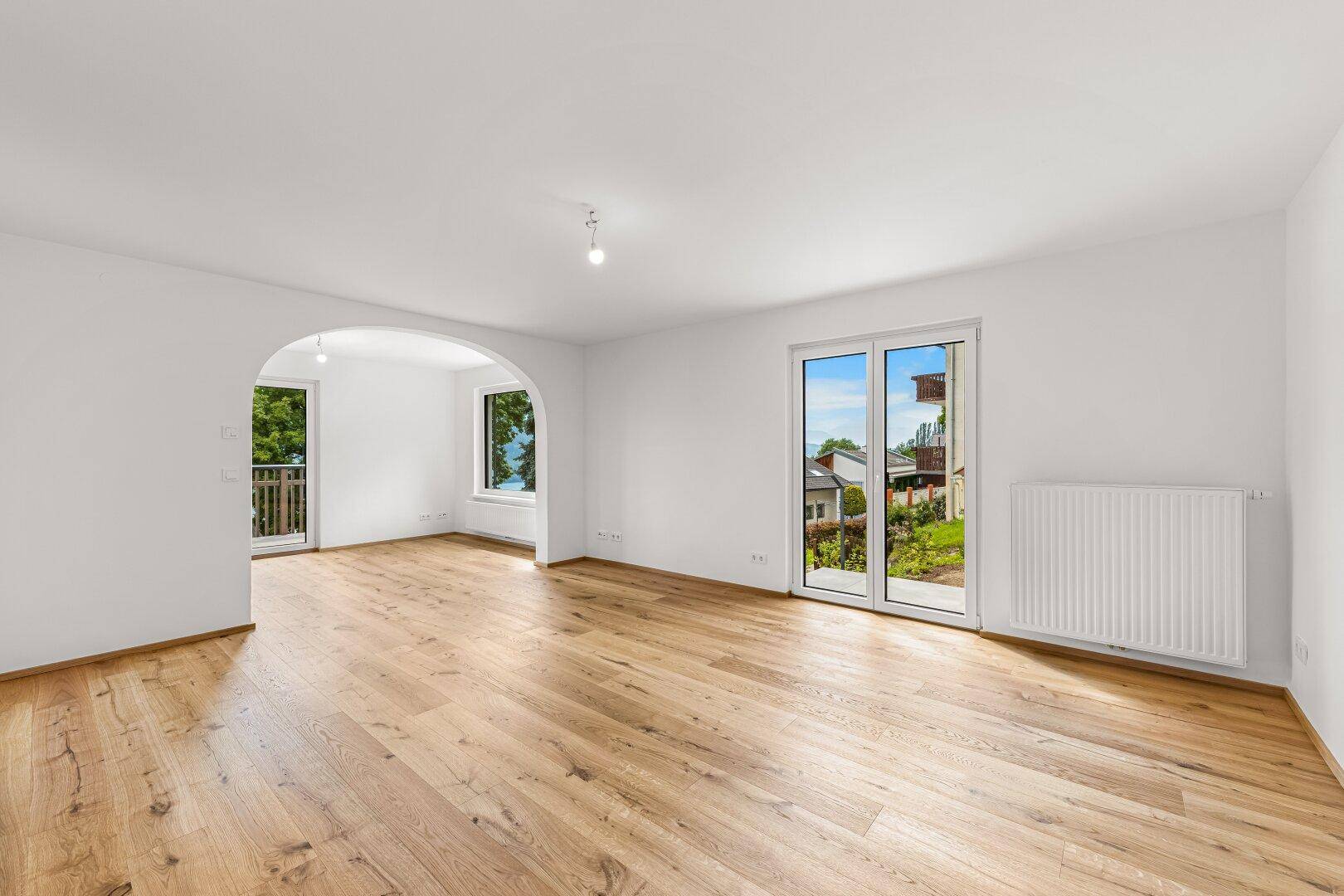 Helles, leeres Wohnzimmer mit Eichenparkett, großen Fenstern und Zugang zum Garten.