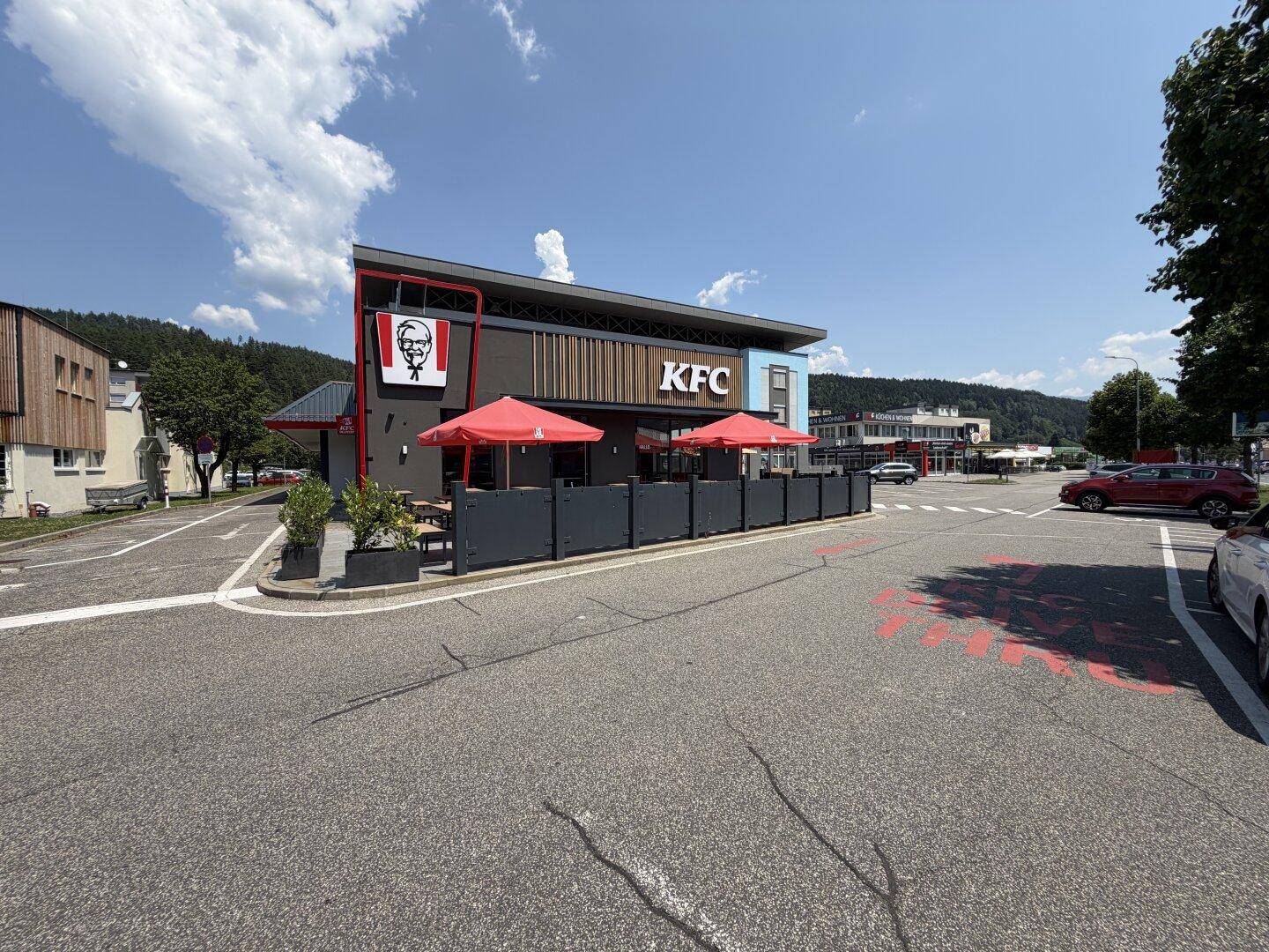 Modernes Fast-Food-Restaurant mit rotem Sonnenschirm und Außenbereich, umgeben von Parkplätzen.