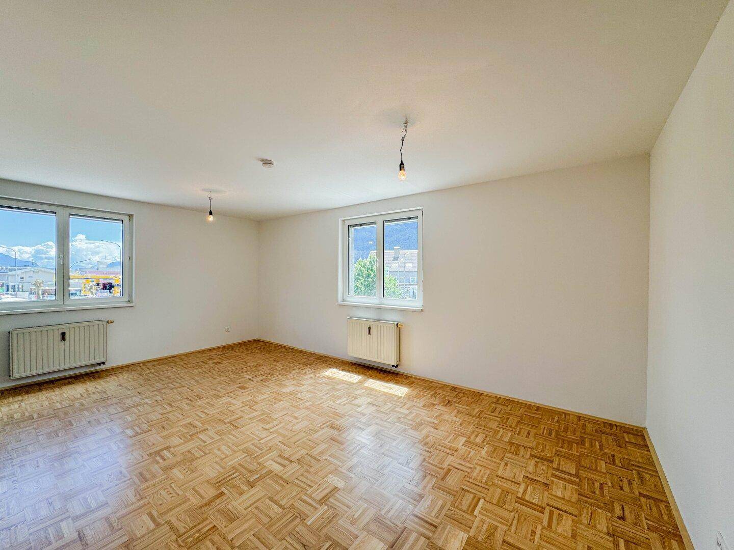 Geräumiger, heller Raum mit Parkettboden und mehreren Fenstern, die viel Tageslicht hereinlassen.