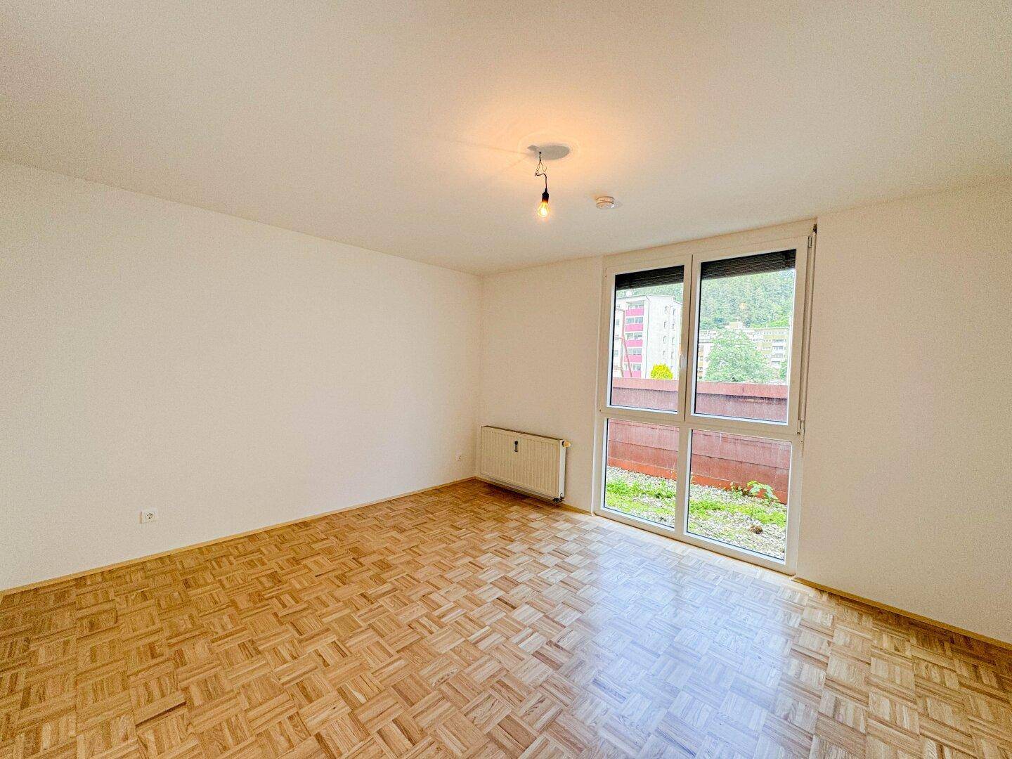Heller Raum mit Parkettboden und großer Fenstertür, die den Blick ins Grüne freigibt.