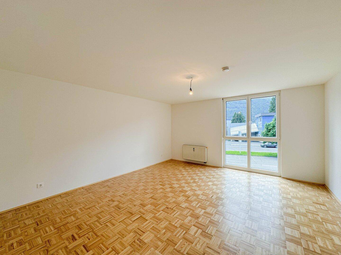 Heller Wohnraum mit Parkettboden und einer großen Fenstertür, die Zugang nach draußen bietet.