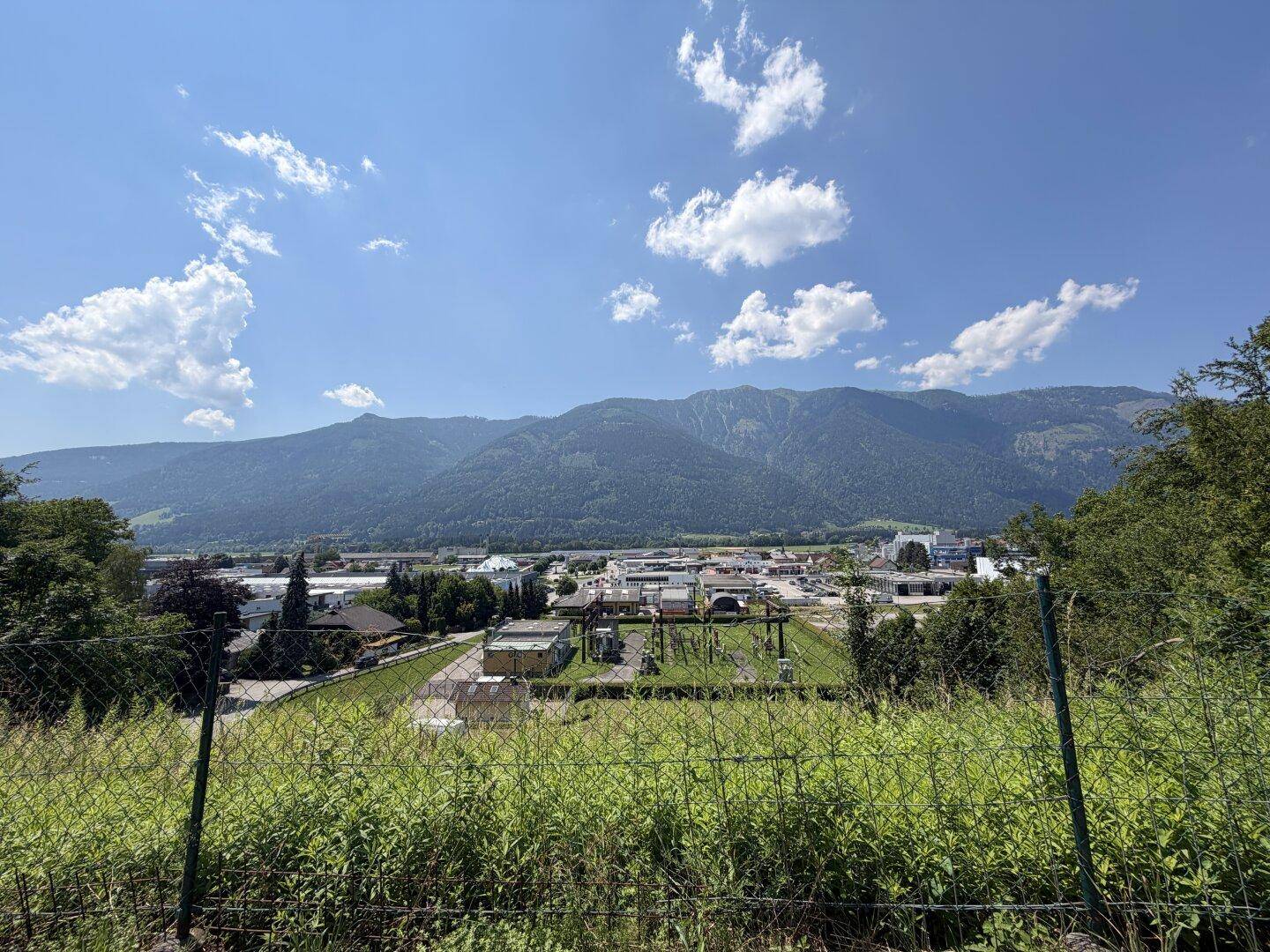 Panoramablick auf eine Stadt und Industrieanlagen im Tal, umgeben von hohen Bergen und blauem Himmel.