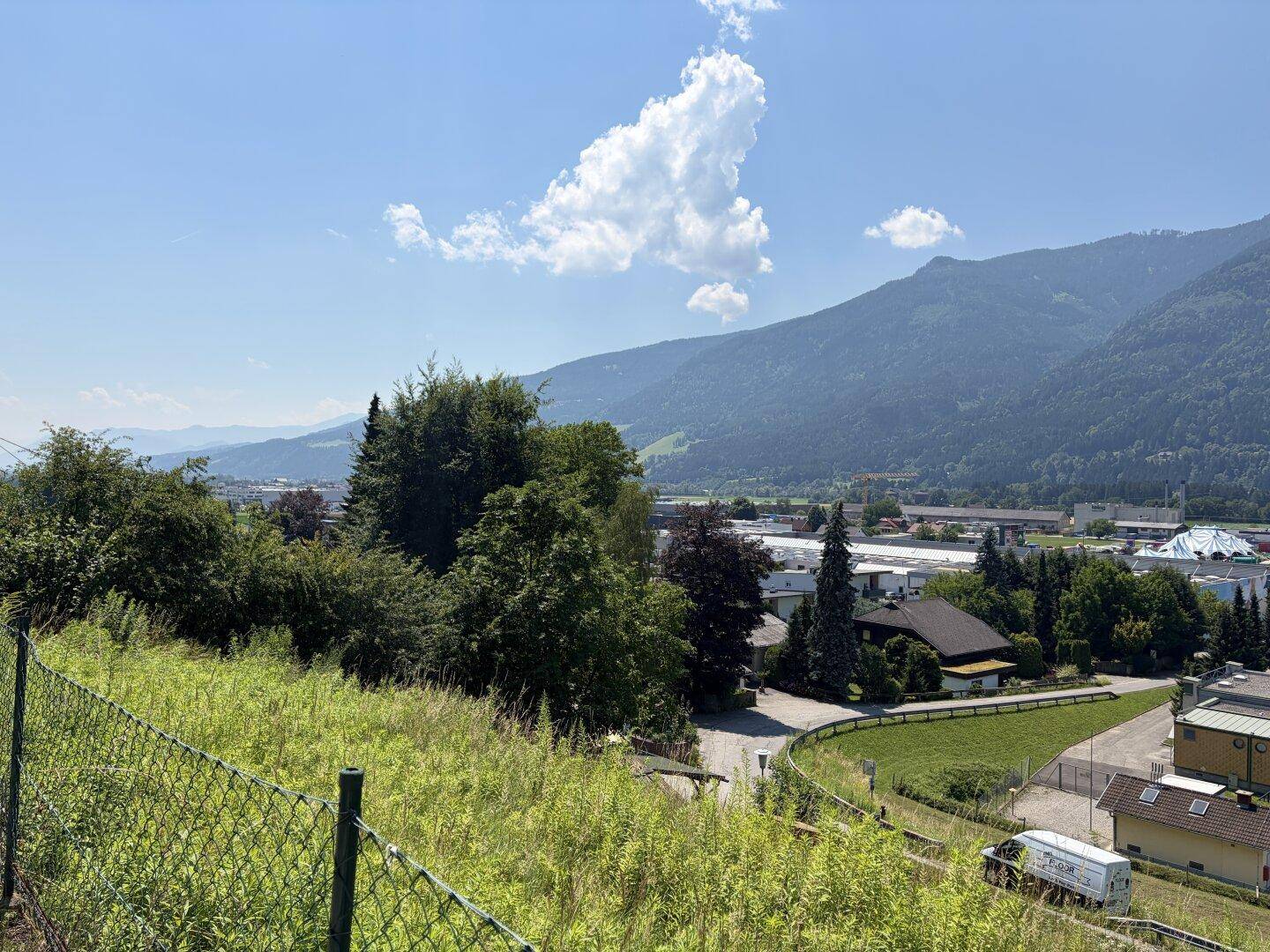 Blick über eine städtische Landschaft im Tal mit Bergen am Horizont, teilweise durch Bäume verdeckt.