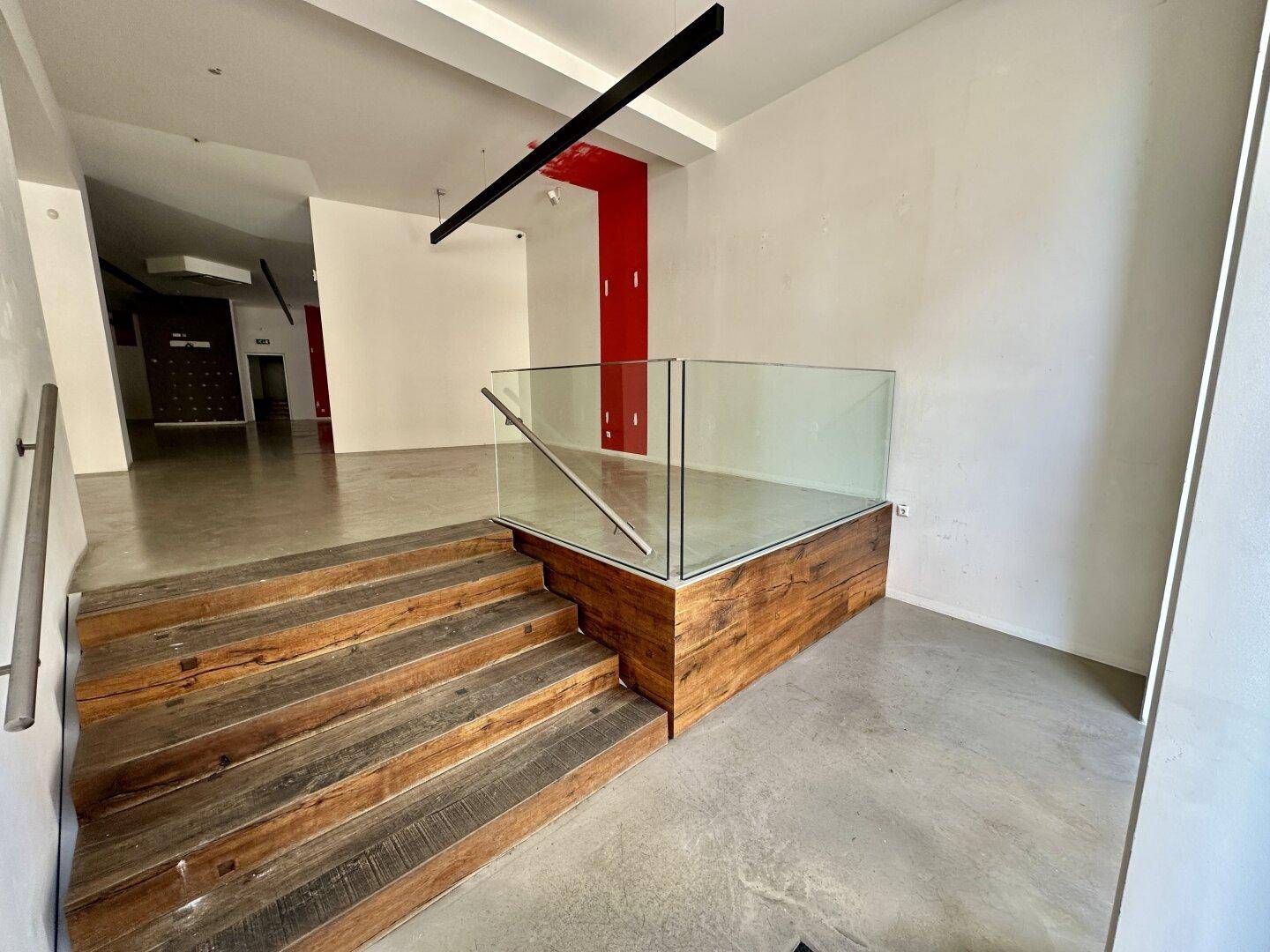 Eingangsbereich eines Geschäfts mit Holztreppe, Glasgeländer und Betonboden.