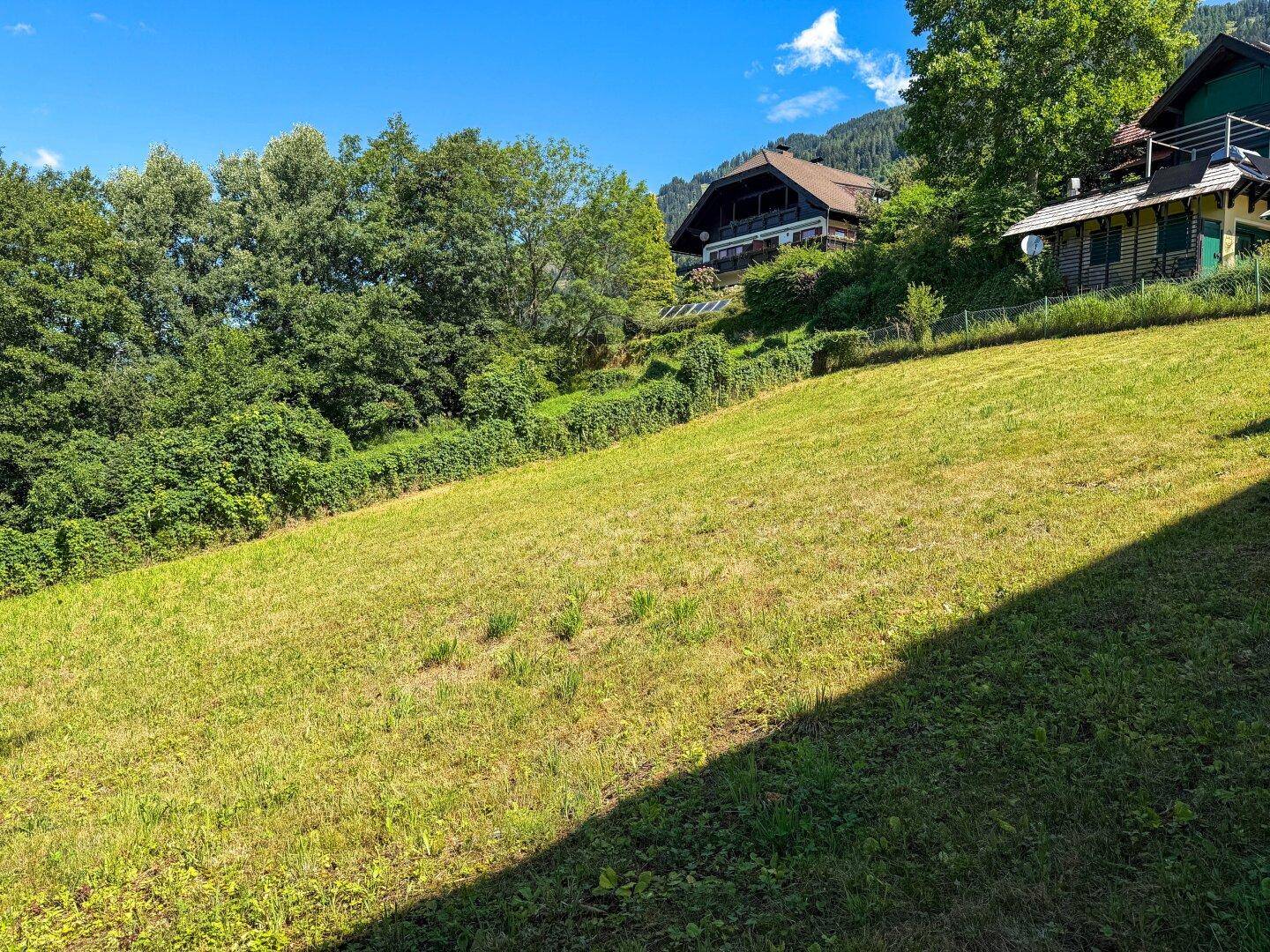 Grüne Hangwiese mit Blick auf traditionelle Häuser und die bewaldete Umgebung unter strahlend blauem Himmel.