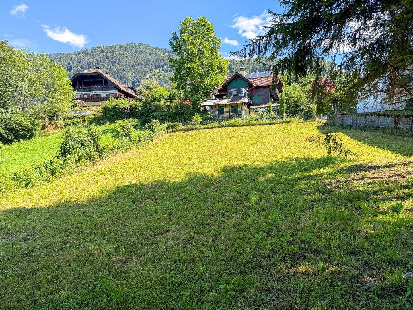 Sonniges Grundstück mit Hanglage, grüner Wiese und Häusern im Hintergrund vor einer Bergkulisse.