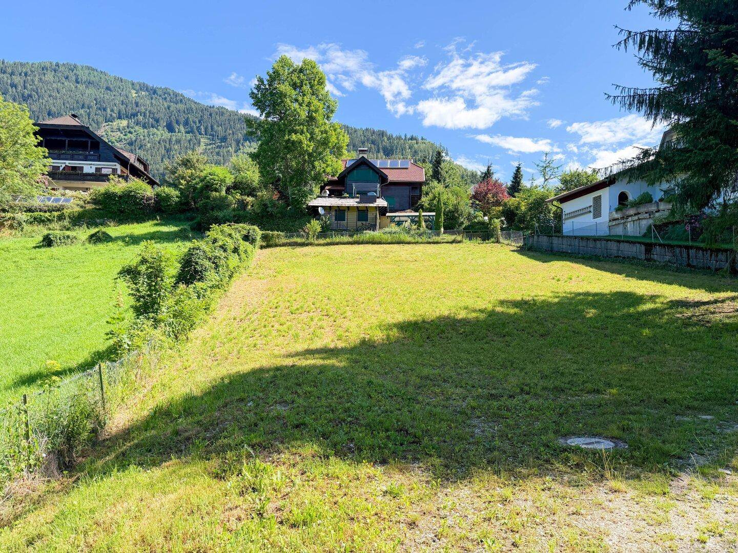 Großes, sonniges Grundstück mit Rasenfläche, umgeben von Hecken und Bäumen, mit Häusern im Hintergrund.