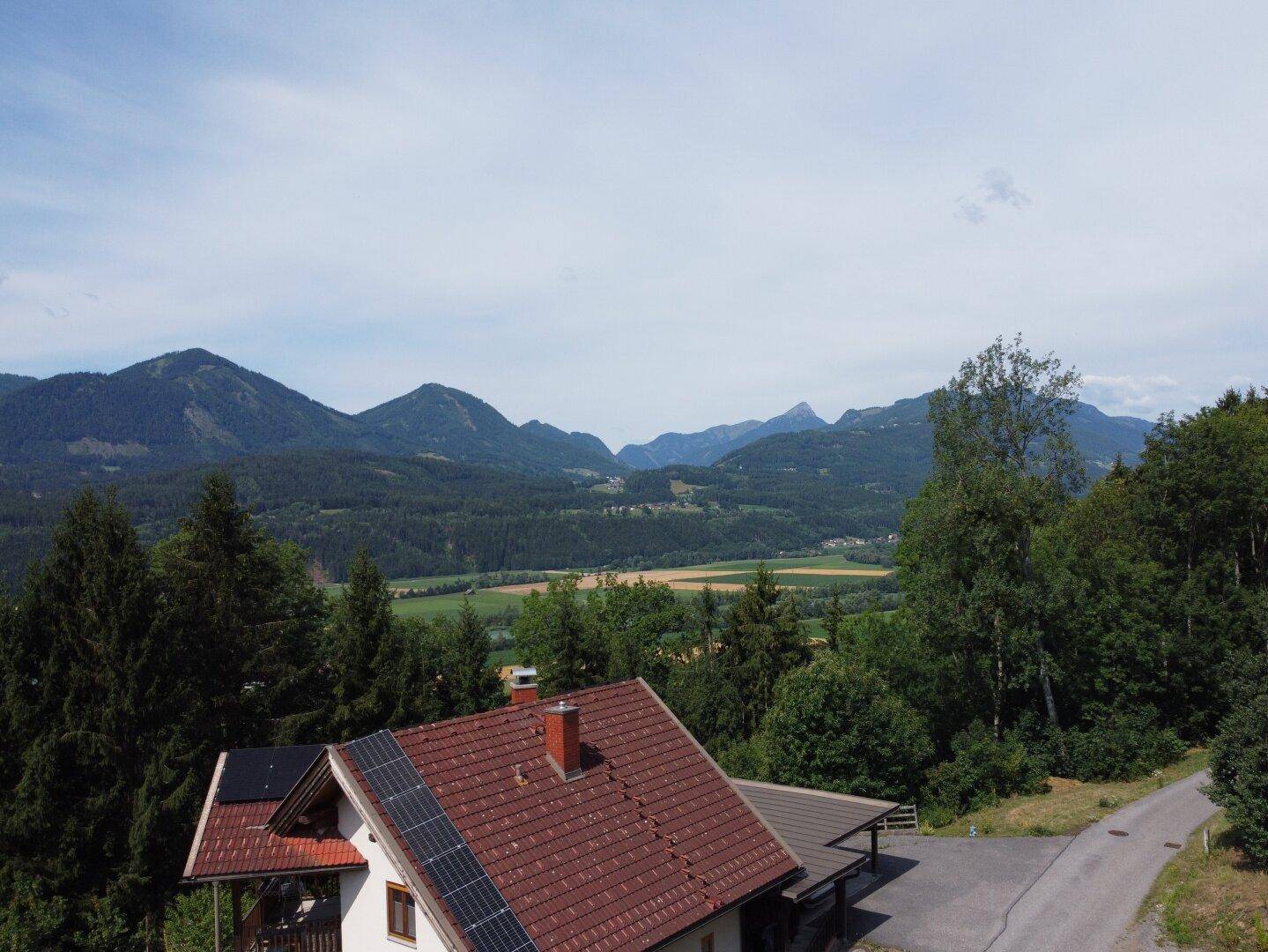 Haus mit Ziegeldach und Solaranlage, umgeben von Bäumen mit Bergblick.