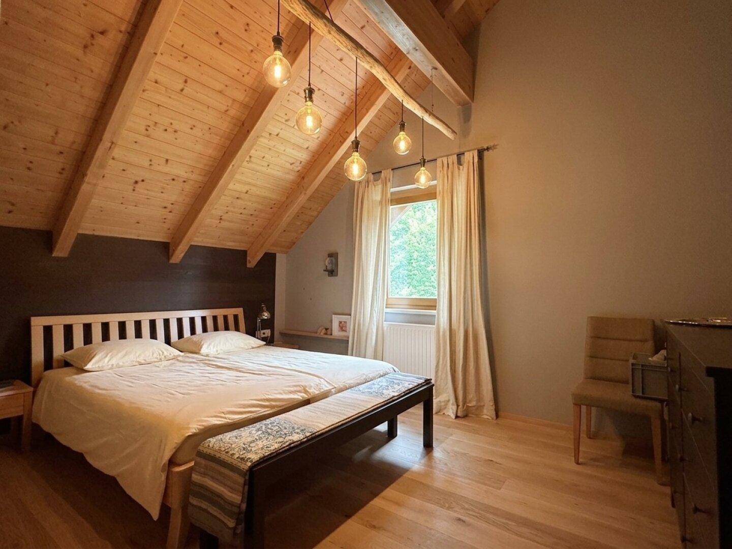 Schlafzimmer mit schräger Holzdecke, Holzboden und natürlichem Lichteinfall durch das Fenster.