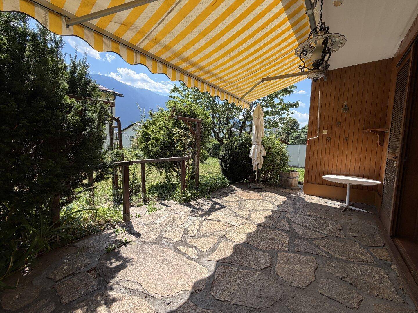 Sonnige Terrasse mit Steinplattenboden, gelb-weiß gestreifter Markise und Bergblick.