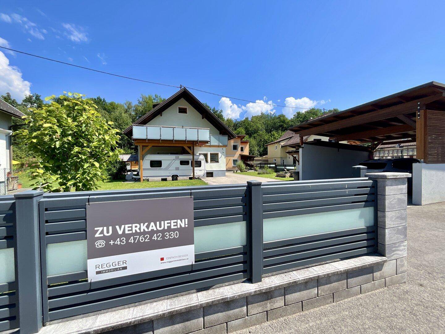 Außenansicht des Hauses mit modernem Zaun, Carport und Verkaufsschild.