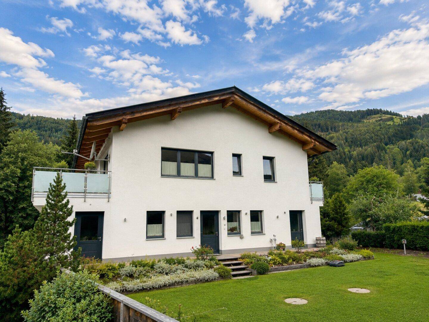 Moderne Hausfassade mit weißem Putz, dunklen Fenstern und Satteldach in alpiner Umgebung.