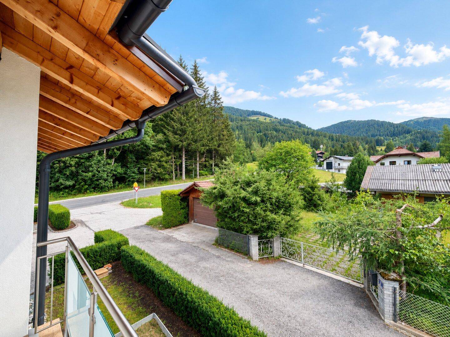 Zufahrt zum Haus mit Hecke und Blick auf die Straße und die bewaldete Berglandschaft.