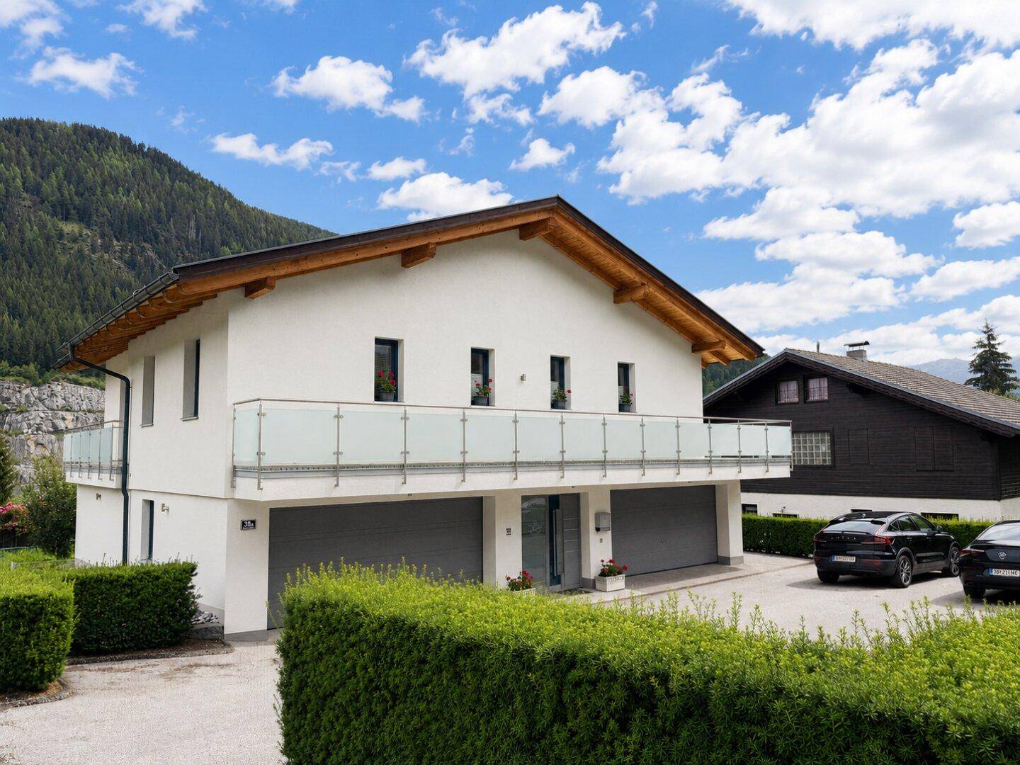Moderne Hausfassade mit zwei Garagen und Balkon, eingebettet in eine grüne Umgebung.