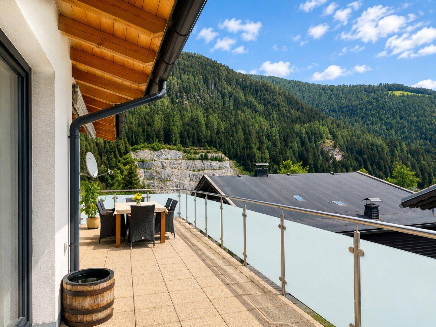 Lange Terrasse mit Fliesenboden und Glasgeländer, umgeben von grünen Bergen.