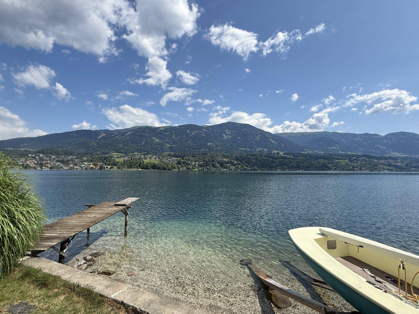 Malerischer Blick auf den See mit Holzsteg, kleinem Boot am Ufer und Bergen im Hintergrund.