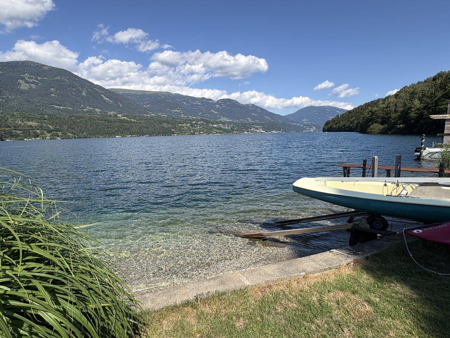 Weitläufiger Blick auf den klaren See mit Boot am Ufer und bewaldeten Bergen im Hintergrund.
